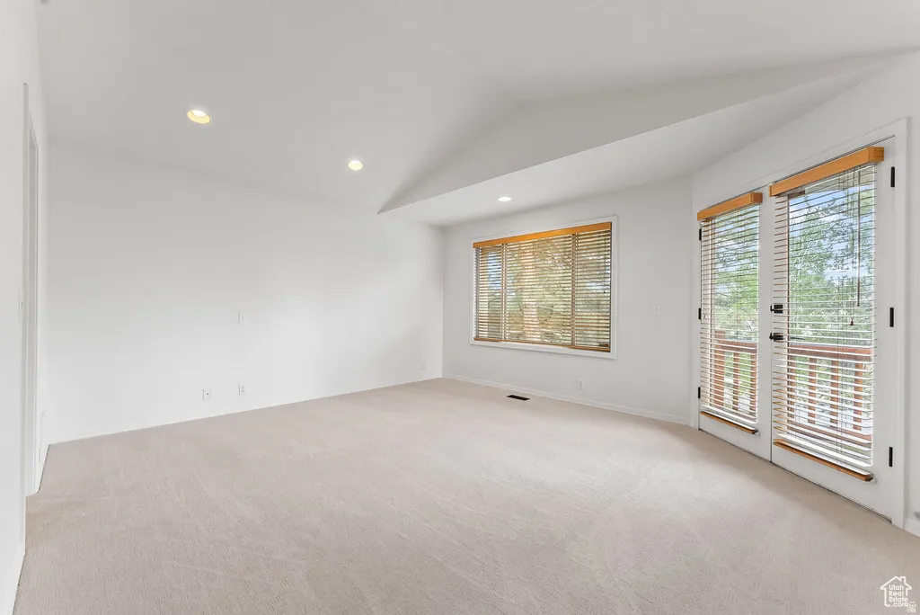 Vaulted ceiling room featuring light-toned carpeting, recessed lighting, a large window with wood blinds, and glass-paned double doors leading to an outdoor area with wooden railings