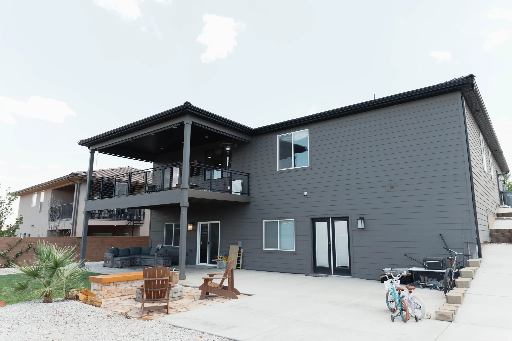 Rear exterior featuring a covered upper-level deck with glass railings, a lower-level concrete patio, and a stone fire pit area