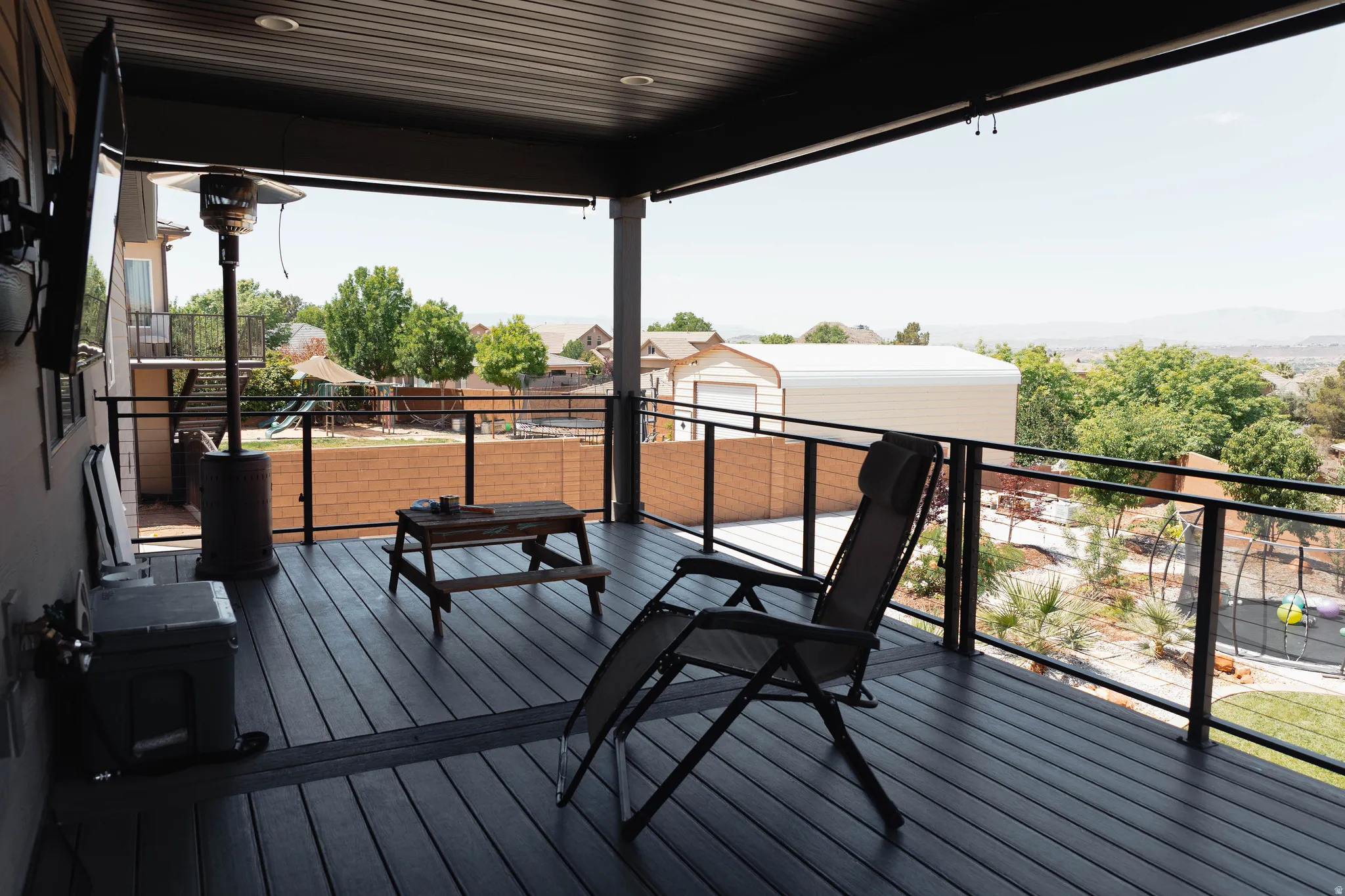 Expansive covered deck featuring composite decking, glass panel railing, recessed lighting, and an exterior wall-mounted television
