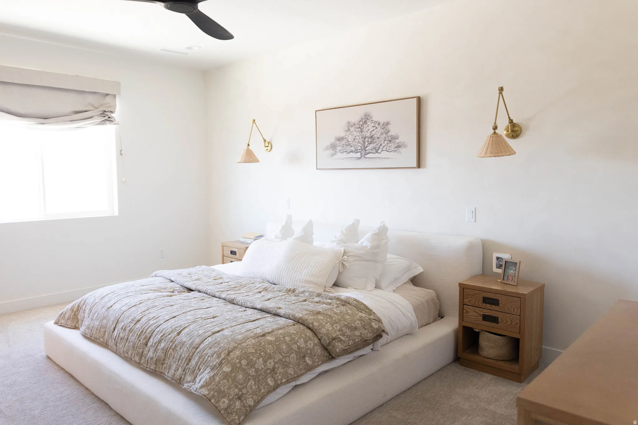 White walls frame this bedroom featuring a ceiling fan, a window with a roman shade, and two wall-mounted sconces