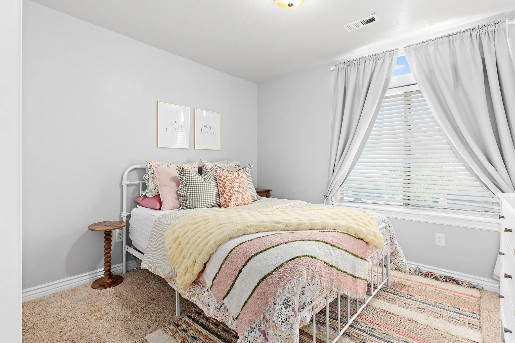 Bright bedroom featuring a large window with blinds and drapes, neutral wall paint, and soft carpet flooring
