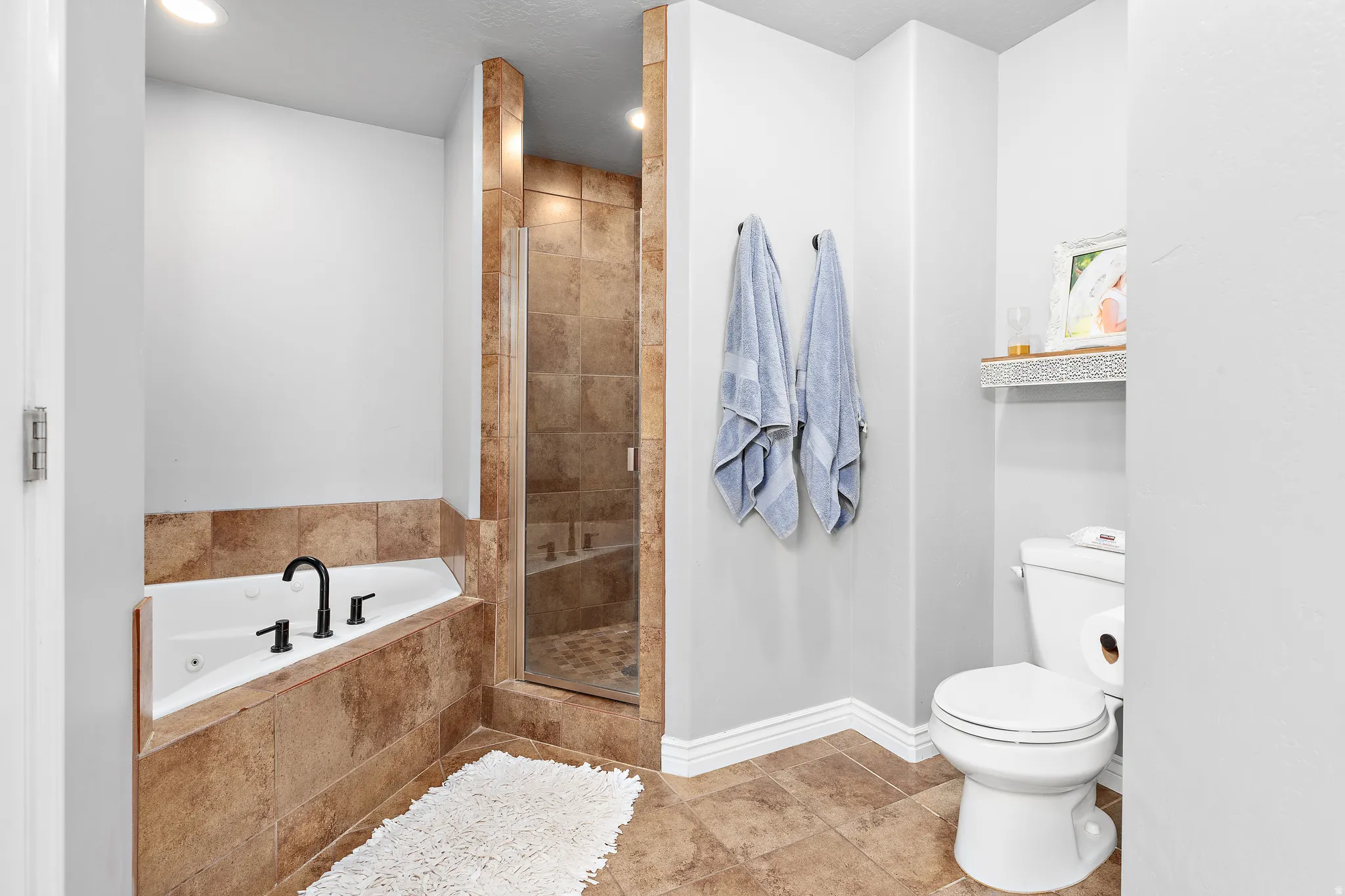 Corner soaking tub with dark bronze fixtures, glass-enclosed walk-in shower with tiled walls and flooring, recessed ceiling lighting, and tiled flooring throughout