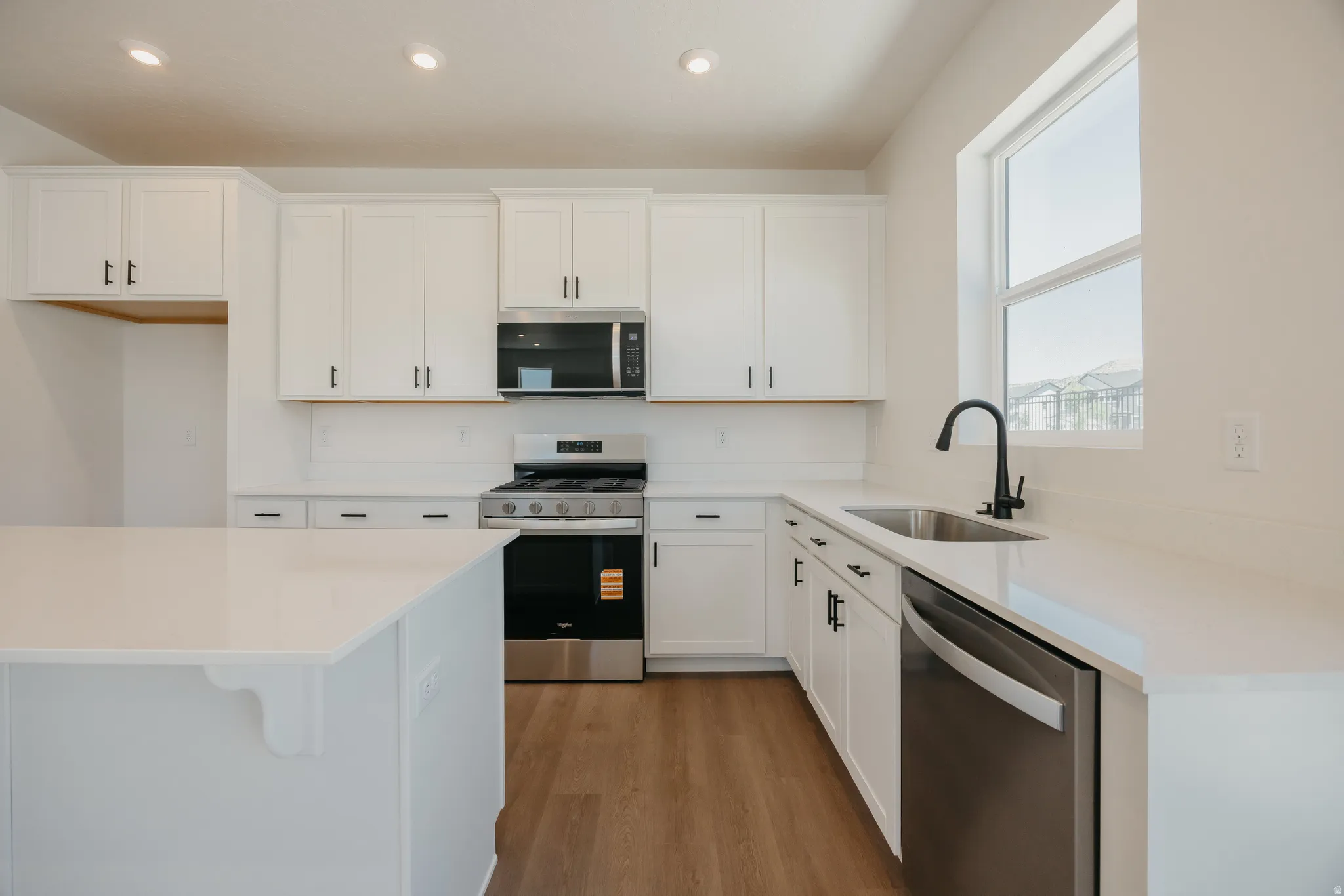 Bright kitchen featuring an island with a solid surface countertop, white cabinetry with black hardware, stainless steel appliances, and wood-finish flooring