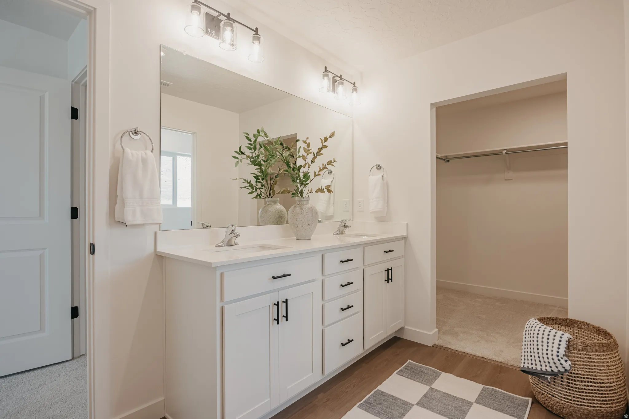 Dual vanity with white cabinetry and dark hardware, large wall-mounted mirror, and wood-finish flooring