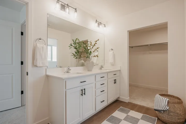 Dual vanity with white cabinetry and dark hardware, large wall-mounted mirror, and wood-finish flooring