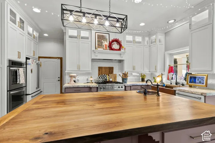 Kitchen featuring a large wood-finish island with integrated sink, white cabinetry with gold-tone hardware, stainless steel double ovens, and a tile backsplash