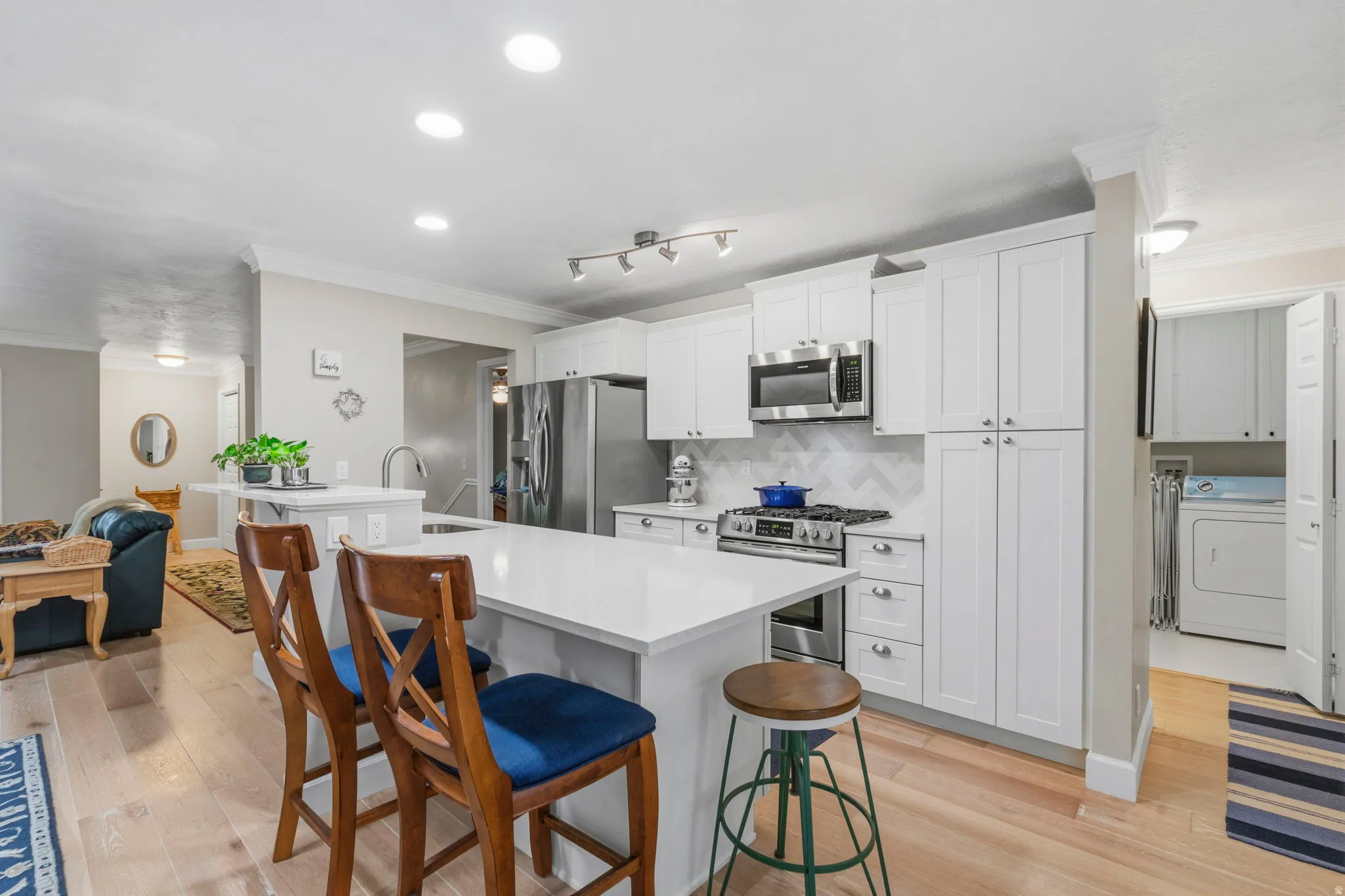 Open-concept kitchen featuring a large peninsula with white/gray speckled quartz countertops, undermount stainless sink, white shaker cabinetry with soft-close doors and drawers, stainless appliances including gas stove/oven, and white oak floors