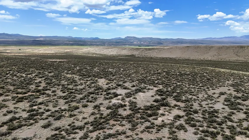 Expansive desert landscape featuring scrub brush, distant mountains, and a clear blue sky with white clouds