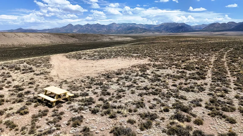 Expansive desert landscape featuring natural brush, a light-colored picnic table, and distant mountain ranges under a partly cloudy sky