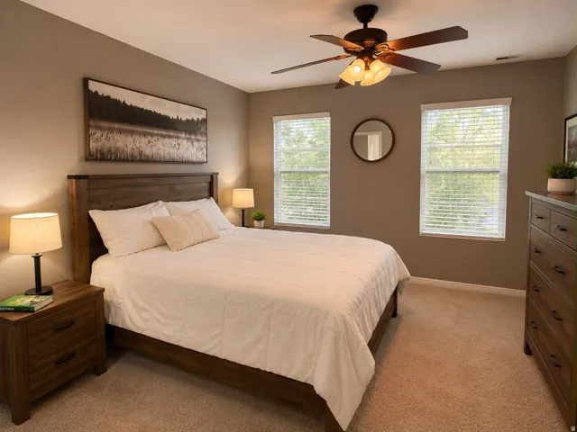 Carpeted room featuring a wood-finish bed frame, two windows with blinds, a ceiling fan with integrated lighting, and a wood-finish dresser