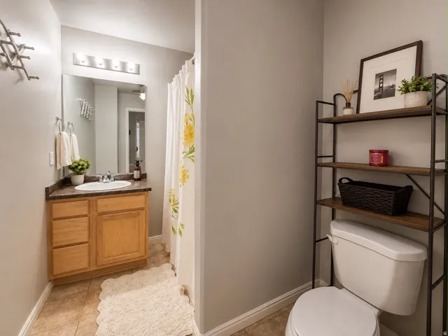 Bathroom featuring a wood-finish vanity with a built-in sink and dark countertop, an overhead mirror with vanity lighting, and tile flooring
