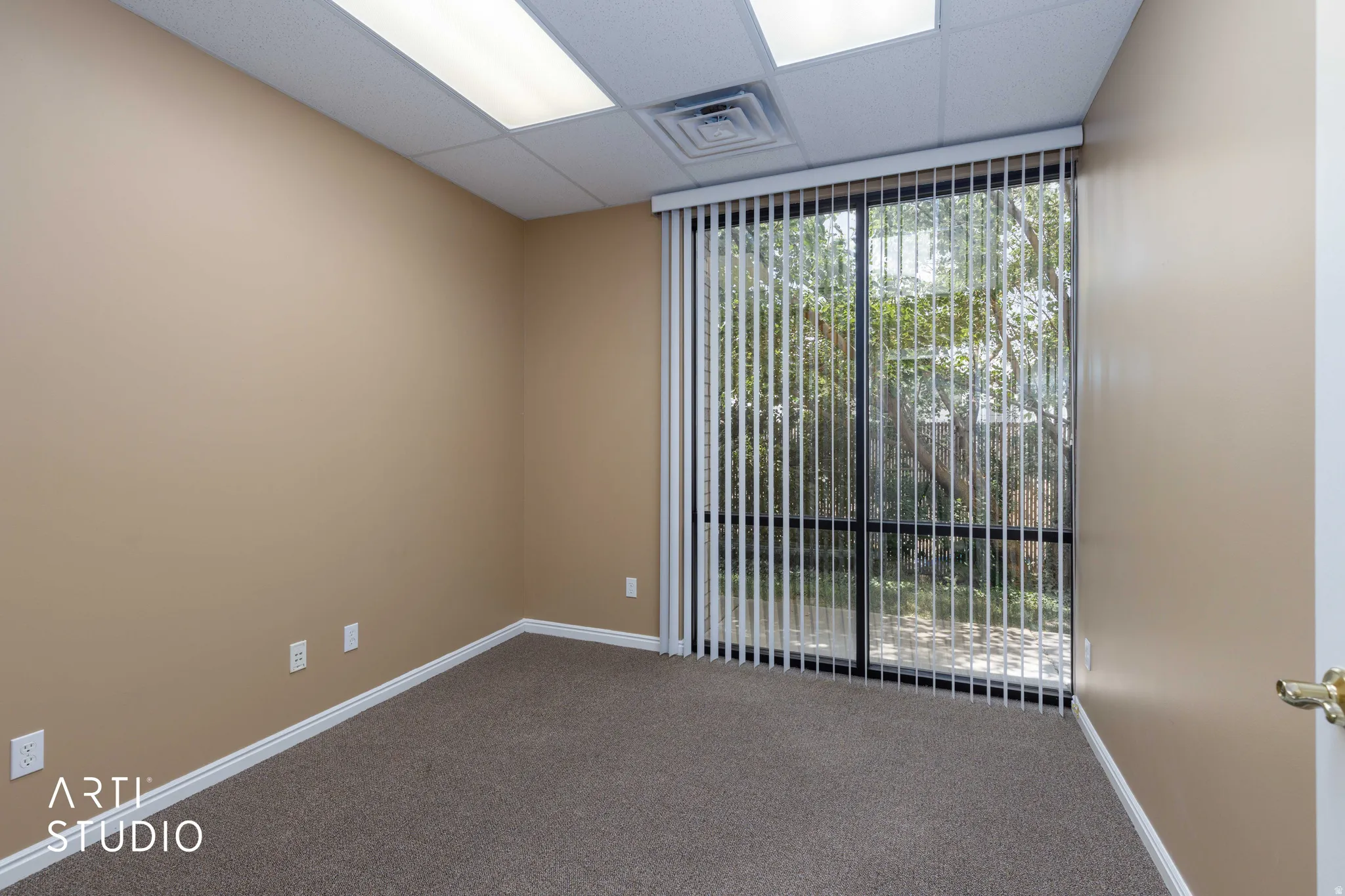 Spacious room featuring neutral carpeting, a drop ceiling with fluorescent lighting, and a large sliding glass door with vertical blinds