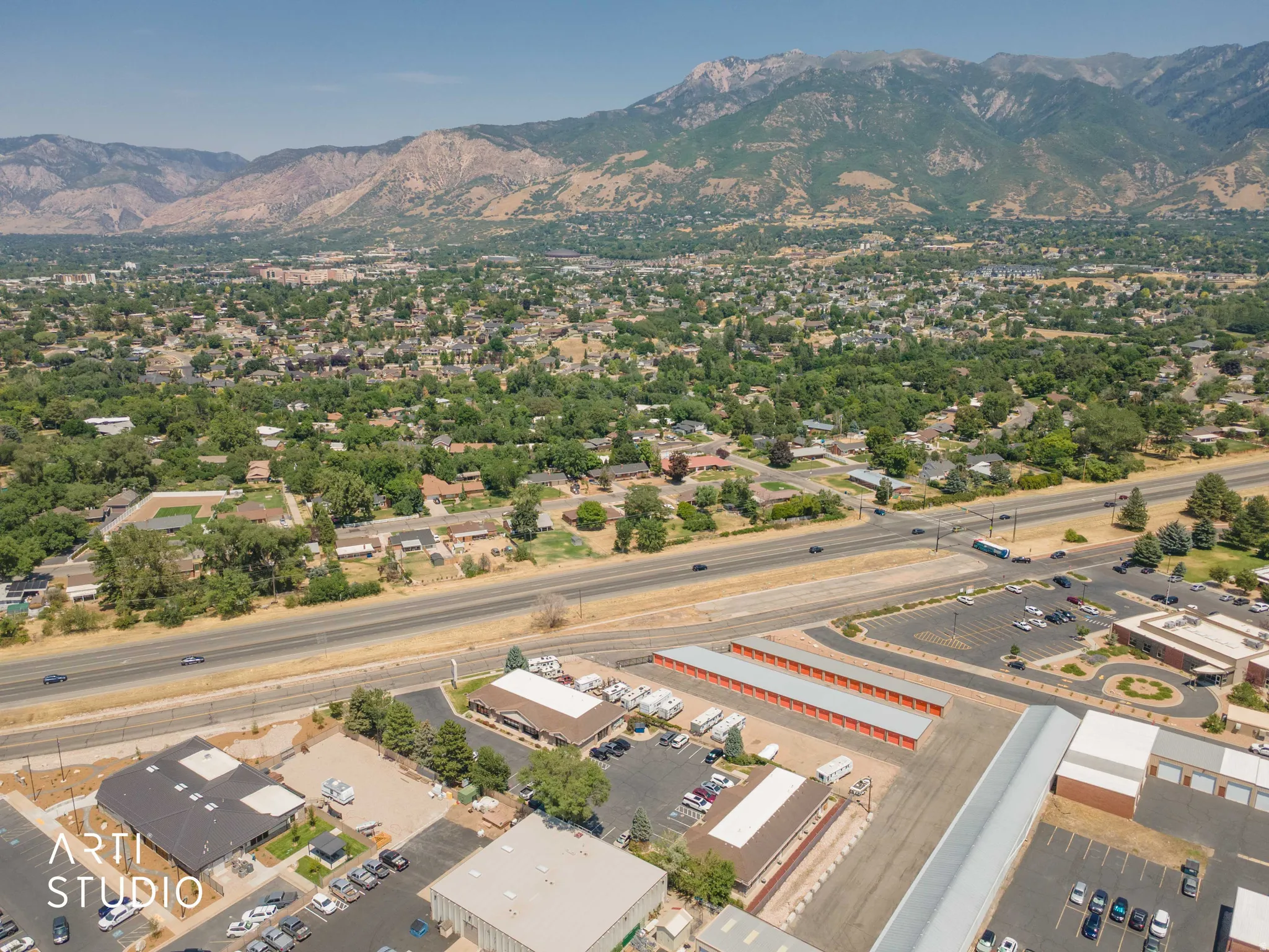Expansive aerial perspective showcasing a mountainous backdrop, a verdant residential area, a multi-lane thoroughfare, commercial structures, and a storage facility