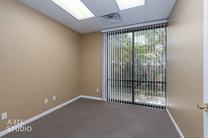 Spacious room featuring neutral carpeting, a drop ceiling with fluorescent lighting, and a large sliding glass door with vertical blinds
