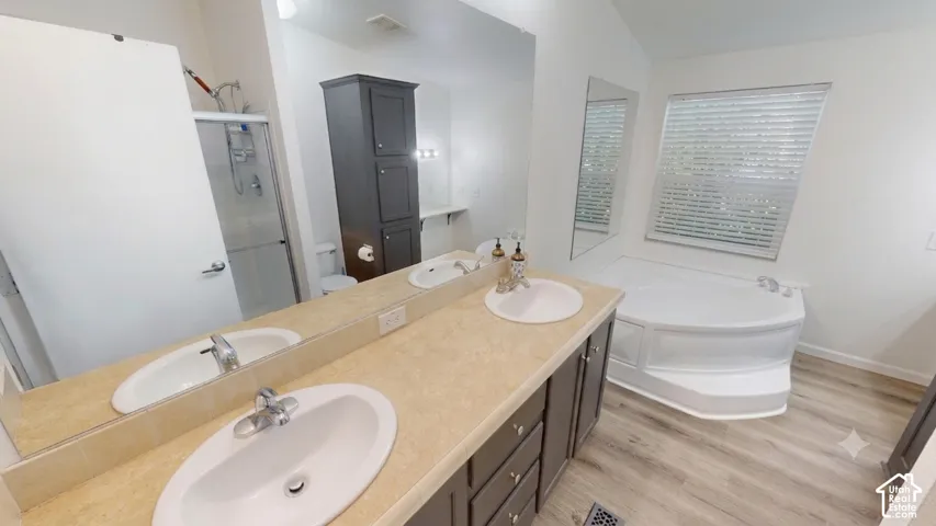 Bathroom featuring a dual vanity with undermount sinks, a large corner soaking tub, a separate glass-enclosed shower, and wood-finish flooring