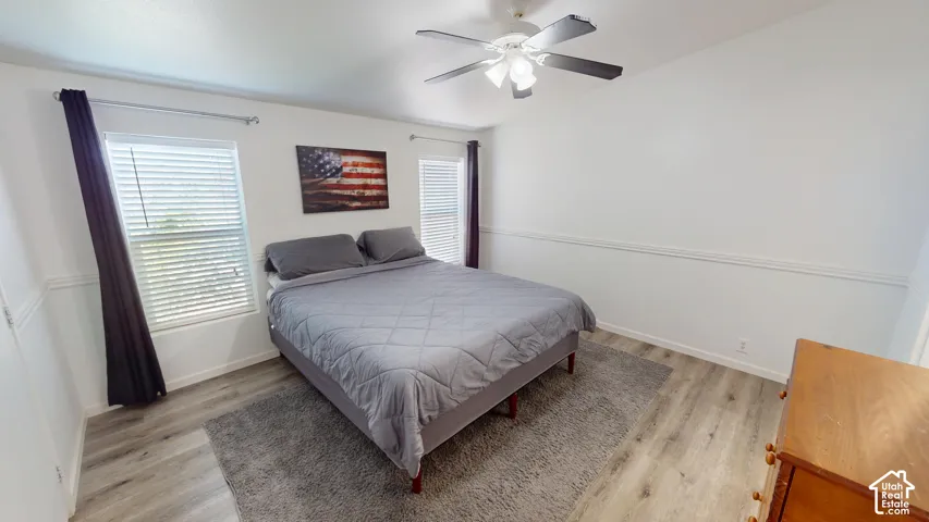 Bedroom featuring wood-finish flooring, two windows with blinds, a ceiling fan with integrated lighting, white walls, and decorative trim