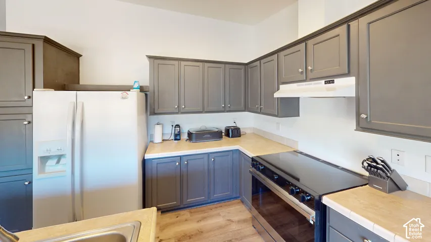 Kitchen featuring grey cabinetry, light-toned countertops, wood-finish flooring, a white side-by-side refrigerator, and a black electric range with an overhead vent