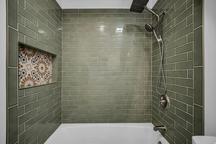 Bathroom featuring a dark gray vanity with a white countertop and integrated sink, wood-finish flooring, a white bathtub with green subway tile surround, and a rectangular mirror with a metal-finish fixture