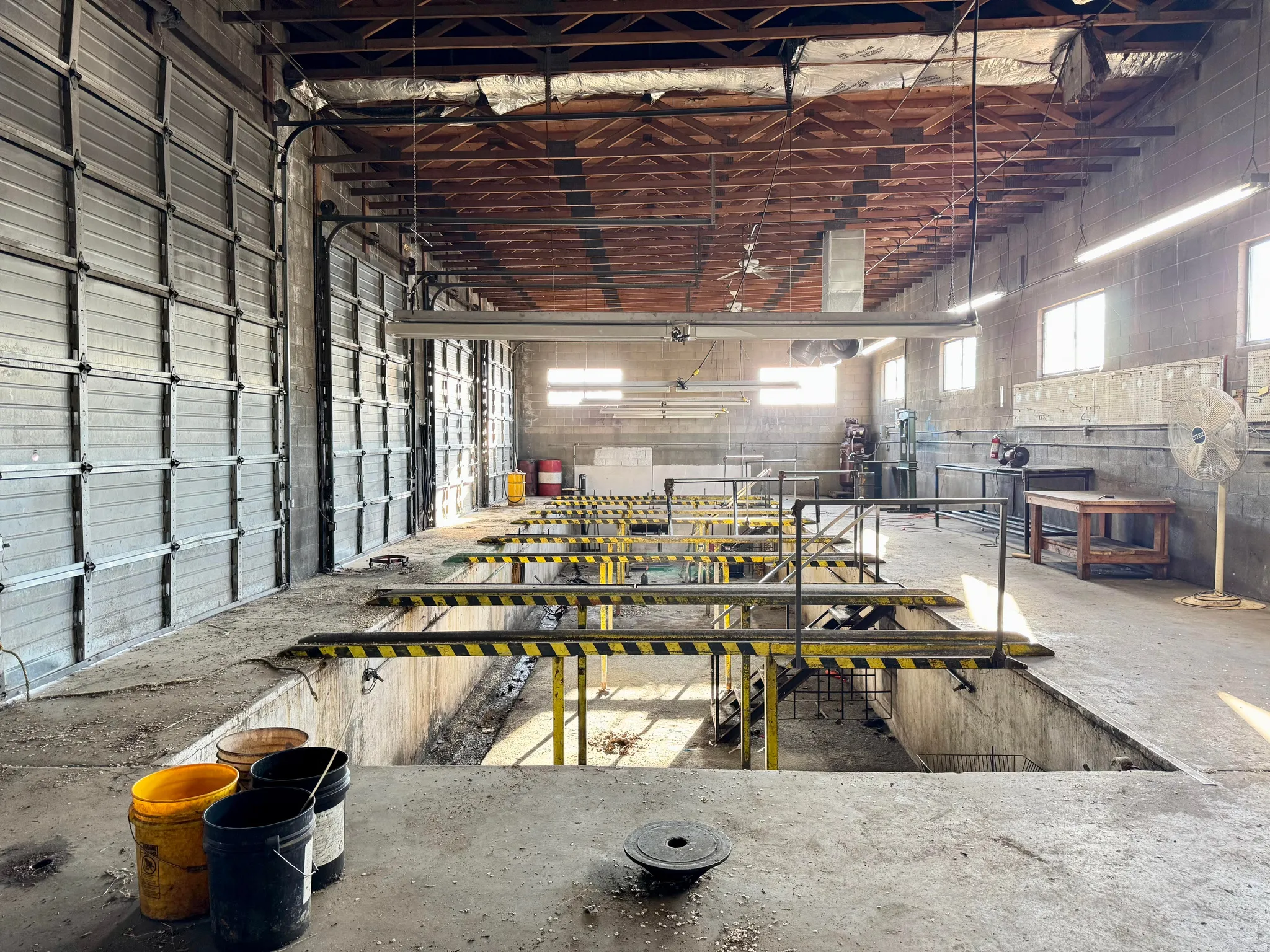 Industrial interior featuring multiple bay doors, exposed wood ceiling beams, concrete flooring, and multiple service pits with safety railings