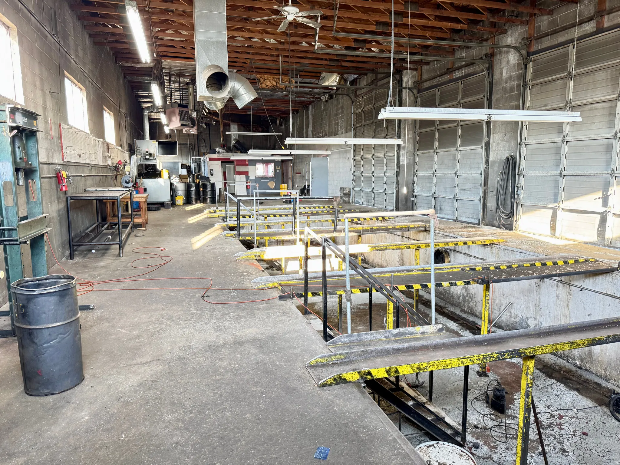Industrial space featuring multiple overhead garage doors, exposed wood beam ceiling, concrete flooring, fluorescent lighting, and a ceiling fan