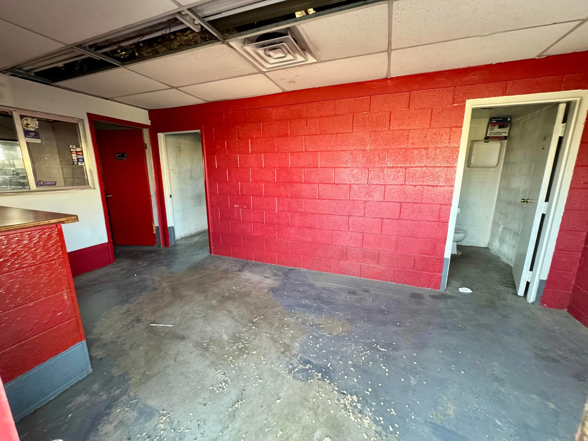 Interior space featuring red painted brick walls, exposed concrete flooring, and a drop ceiling with some missing panels