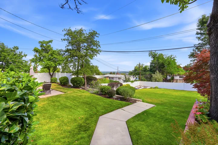 Expansive lawn area featuring a concrete pathway, privacy fencing, mature trees, and landscaped garden beds