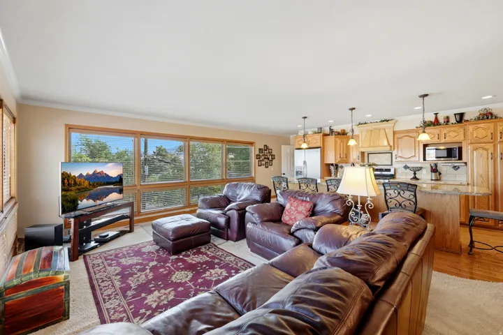 Open-concept living space featuring expansive windows with wood-finish blinds, wood-finish flooring, recessed lighting, and a kitchen with light wood-finish cabinetry, granite countertops, and stainless steel appliances including a built-in microwave and