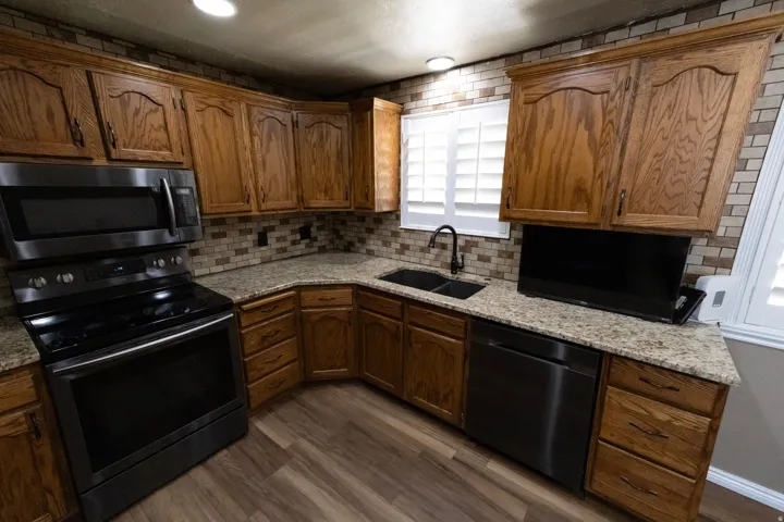 Kitchen featuring wood cabinetry, granite countertops, tile backsplash, wood-finish flooring, and black stainless steel appliances including a range, microwave, and dishwasher