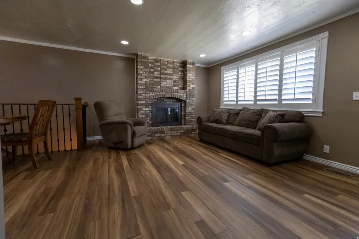 Expansive living area featuring wood-finish flooring, a brick fireplace with a dark insert, recessed ceiling lighting, and a large window with white louvered shutters