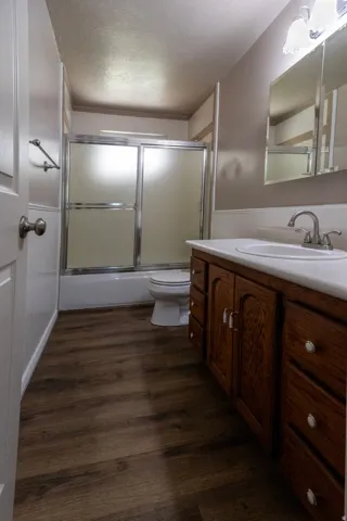 Bathroom featuring wood-finish flooring, a wood-finish vanity with a white countertop and integrated sink, a mirrored medicine cabinet, a shower-tub combination with sliding glass doors, and wainscoting paneling