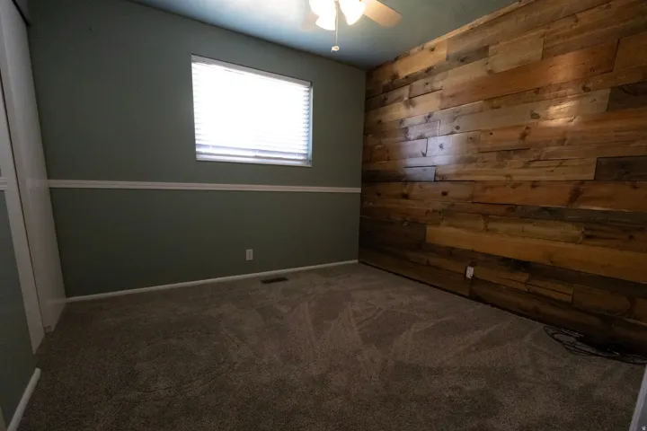 Room featuring a wood-panel accent wall, neutral carpeting, and a ceiling fan