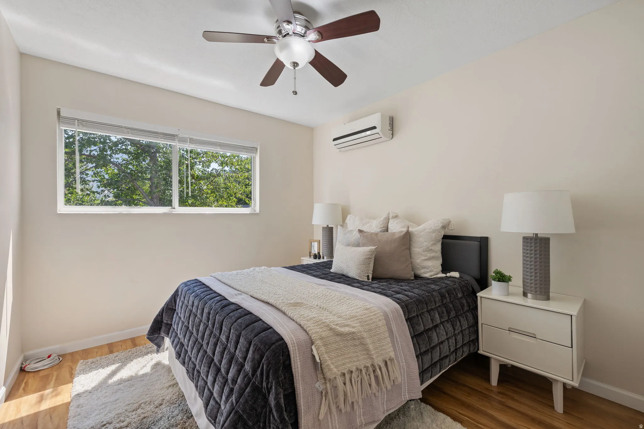 Bedroom featuring wood-finish flooring, a wall-mounted mini-split system, a ceiling fan with integrated lighting, and a large window with privacy blinds