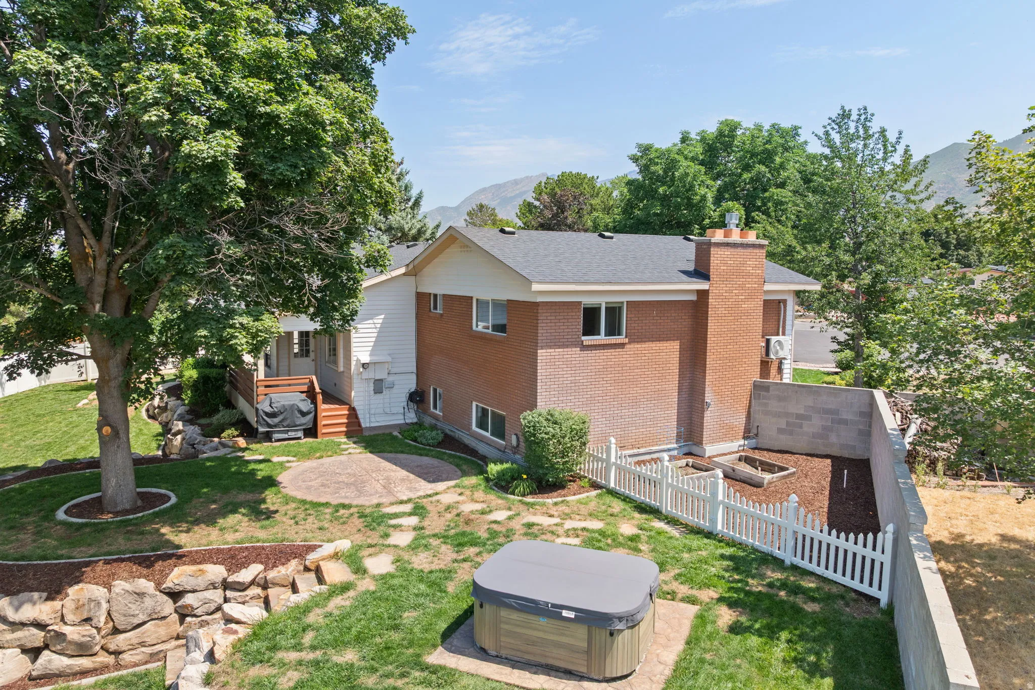 Expansive backyard featuring a large mature tree, a two-story brick and siding exterior, a wood deck with stairs, a stamped concrete patio, and a white picket fence