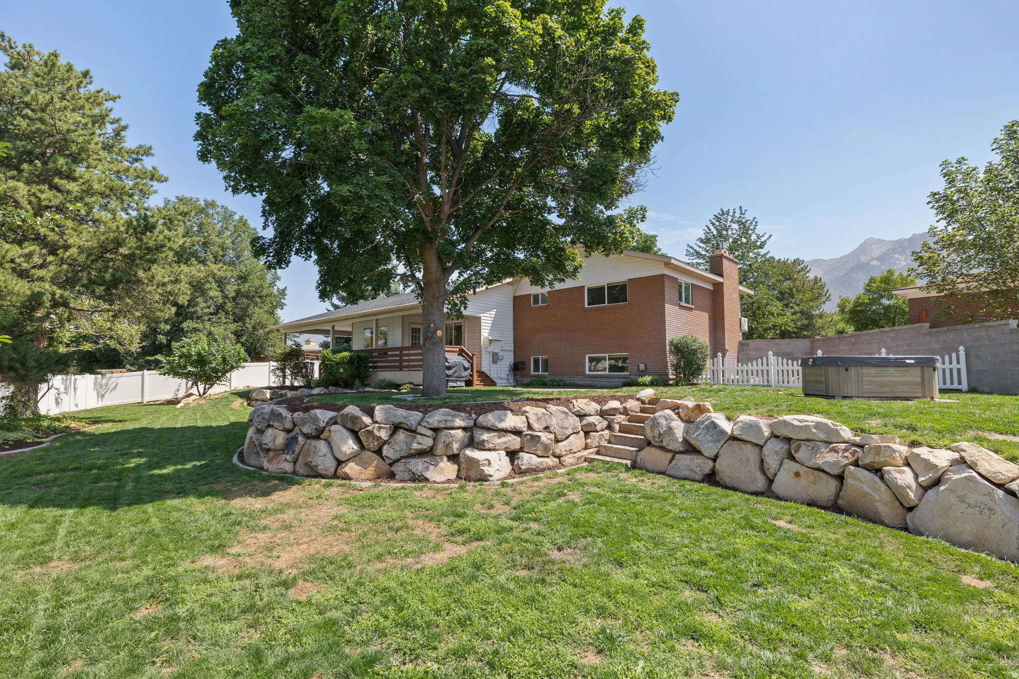 Expansive backyard featuring a tiered landscape with a rock retaining wall, mature trees, and a covered porch
