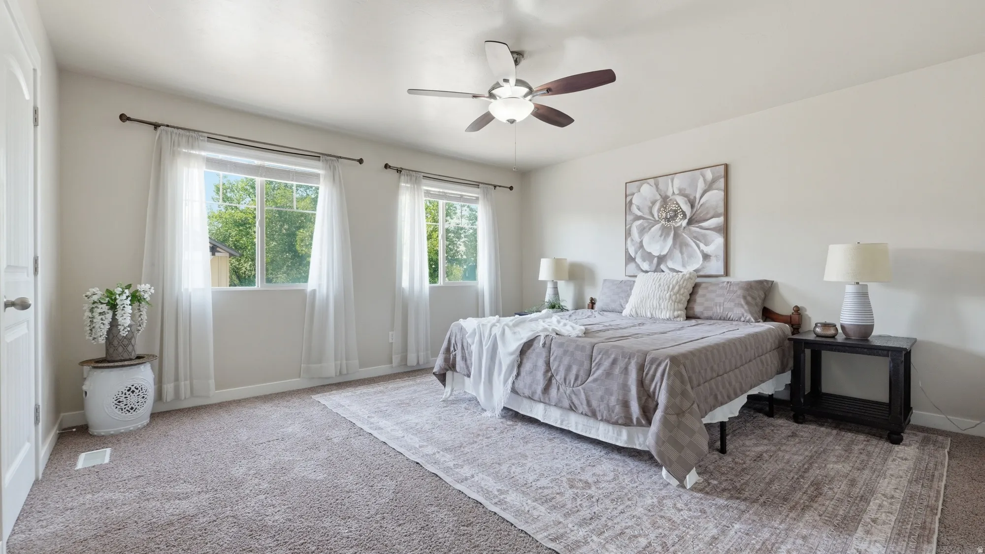 Spacious bedroom featuring neutral carpeting, two large windows, a ceiling fan with integrated lighting, and a solid core door