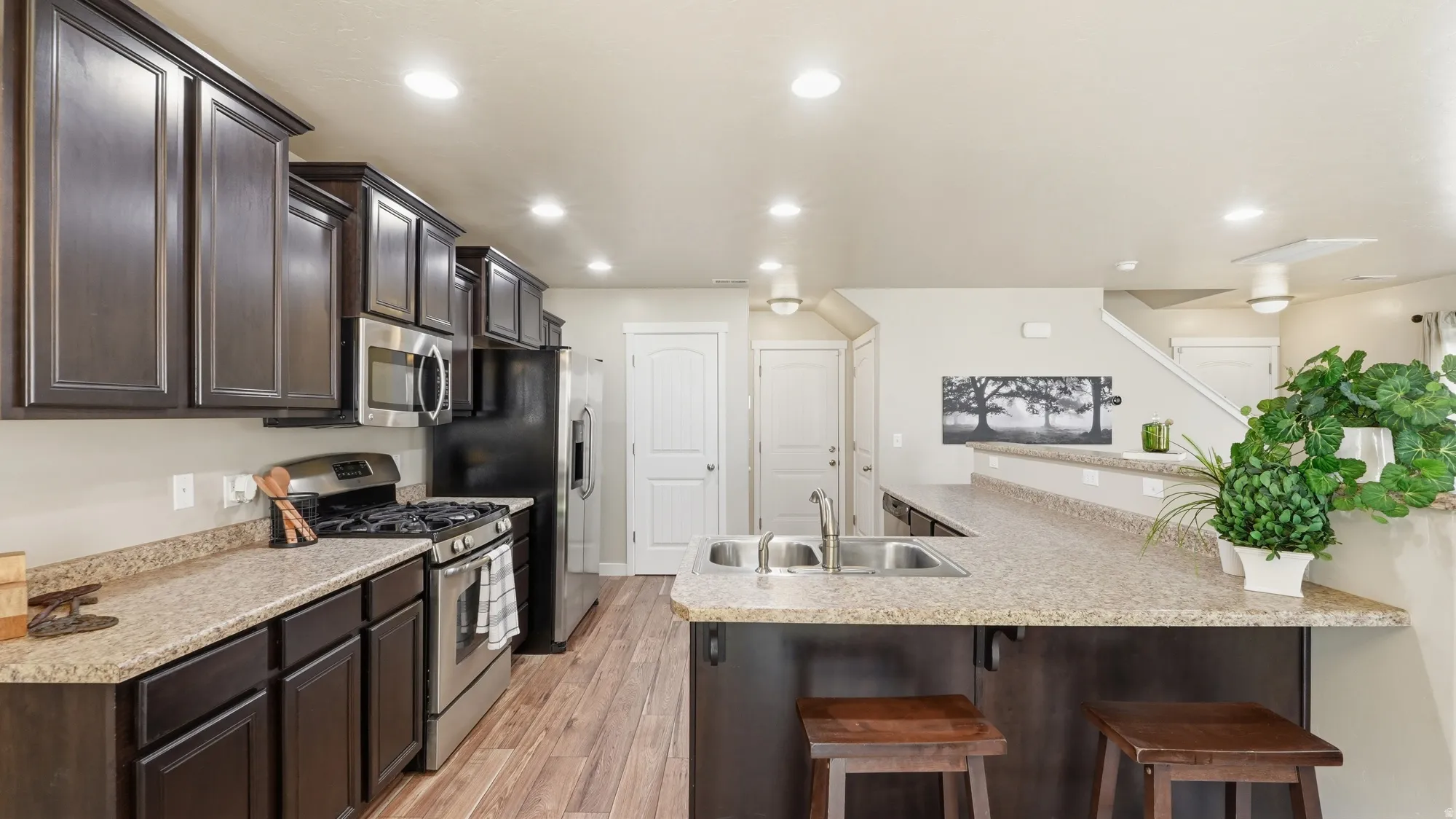 Kitchen featuring dark wood cabinetry, stainless steel appliances, speckled countertops, wood-finish flooring, and a breakfast bar with a double basin sink