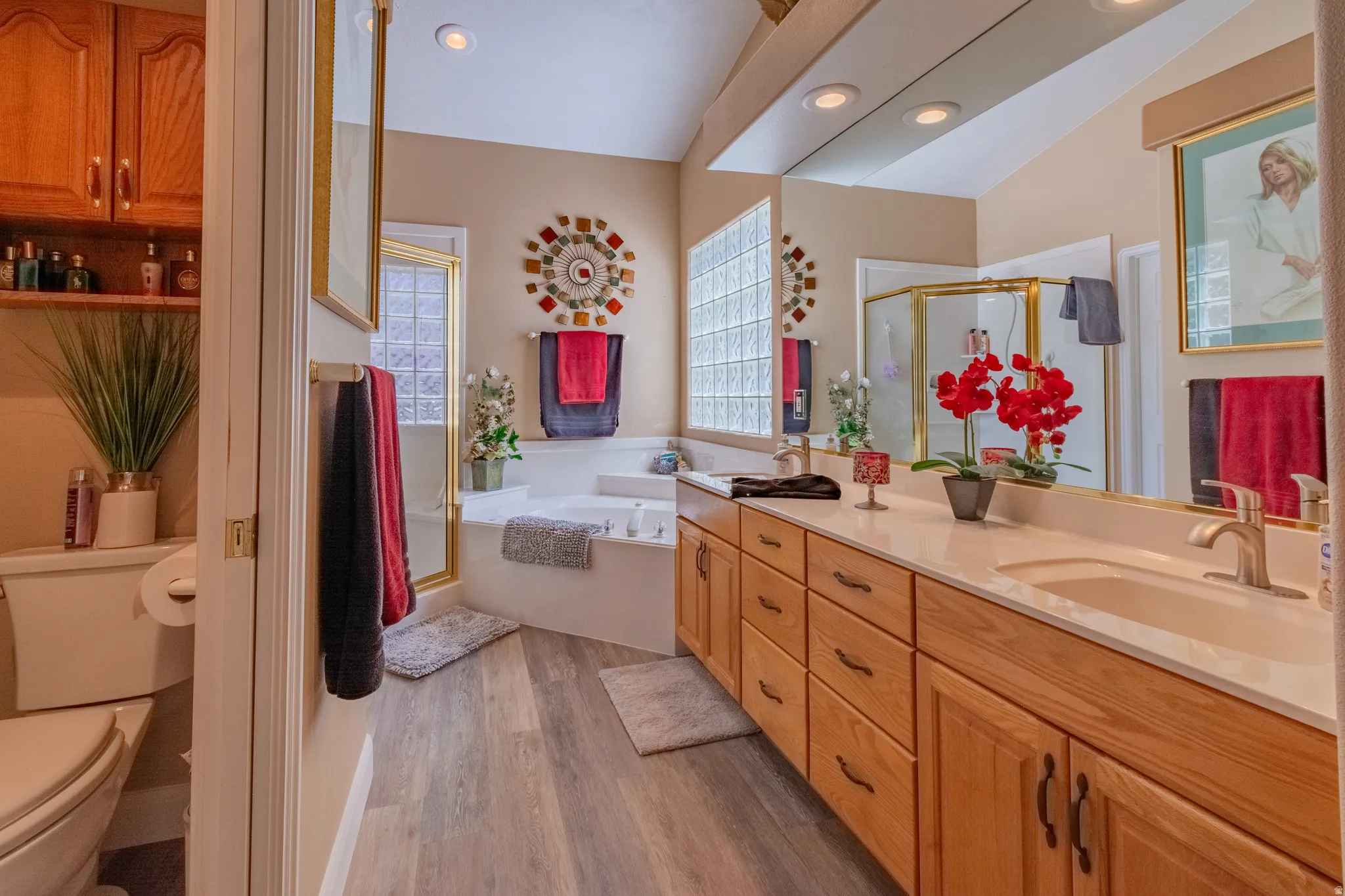 Spacious bathroom featuring a dual vanity with light-colored countertops, a built-in soaking tub, a glass block window, and wood-finish flooring