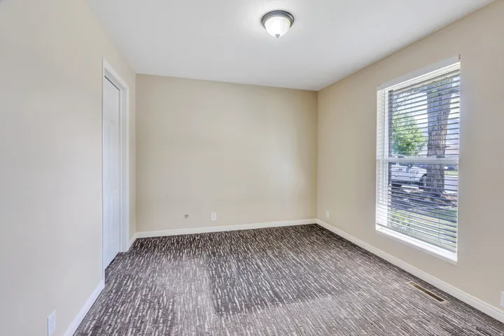 Neutral wall paint, patterned carpeting, natural light from a window with blinds, and a flush-mount ceiling light fixture