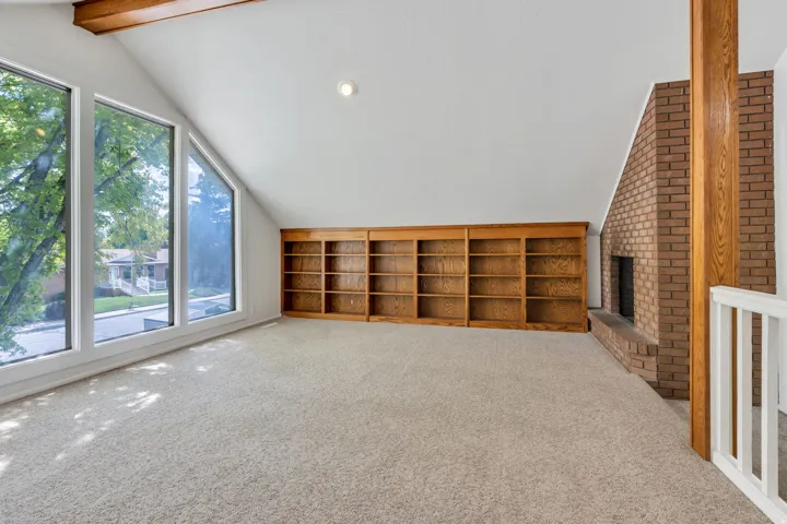 Vaulted ceilings with exposed wood beams, expansive window array, built-in wood shelving, and a brick fireplace