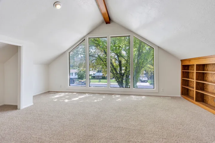 Vaulted ceiling with exposed wood beam, large trapezoidal window, built-in wooden shelving, and neutral carpeting
