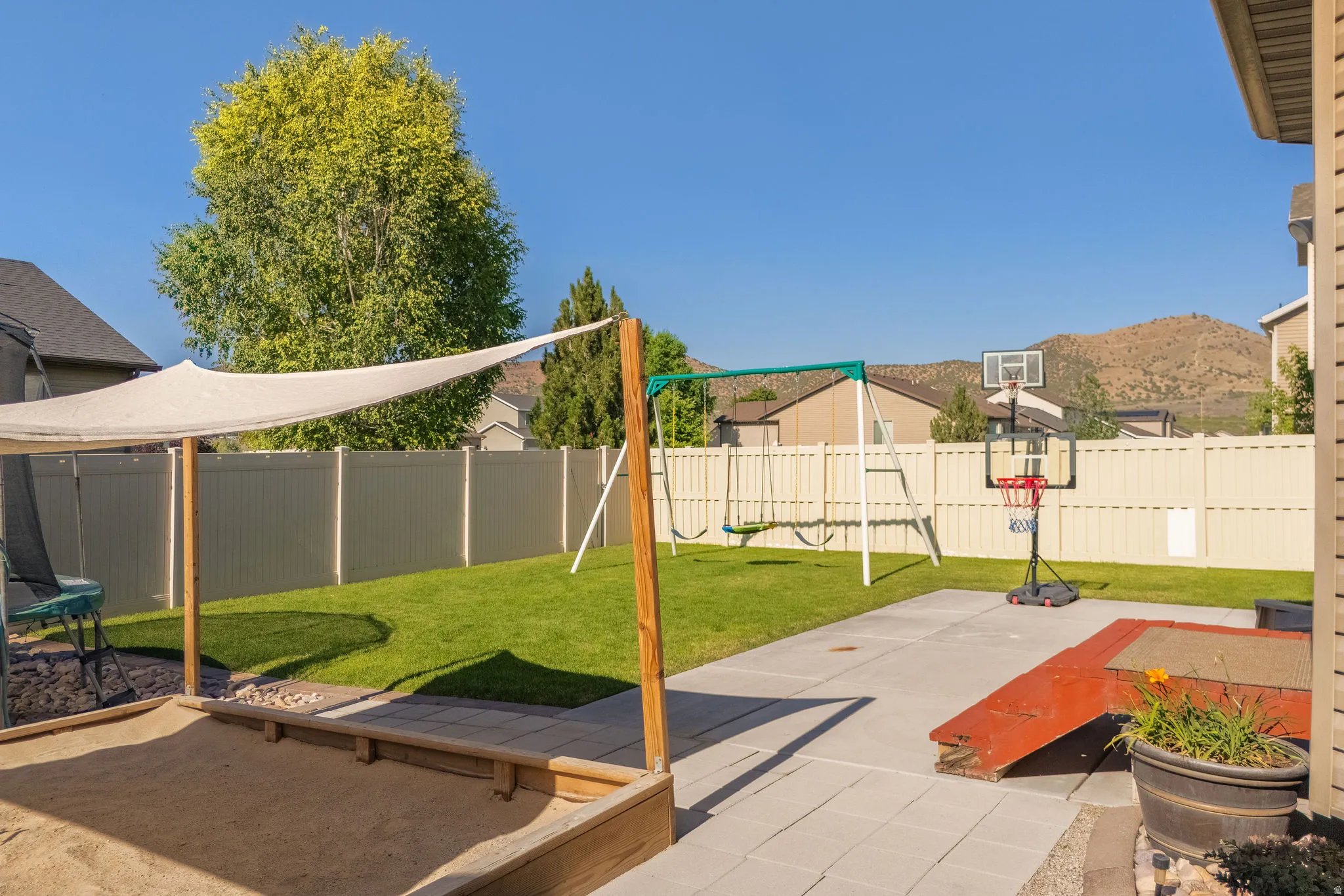 Expansive backyard featuring a manicured lawn, a large sandpit with a sunshade, a swing set, and a paved patio area