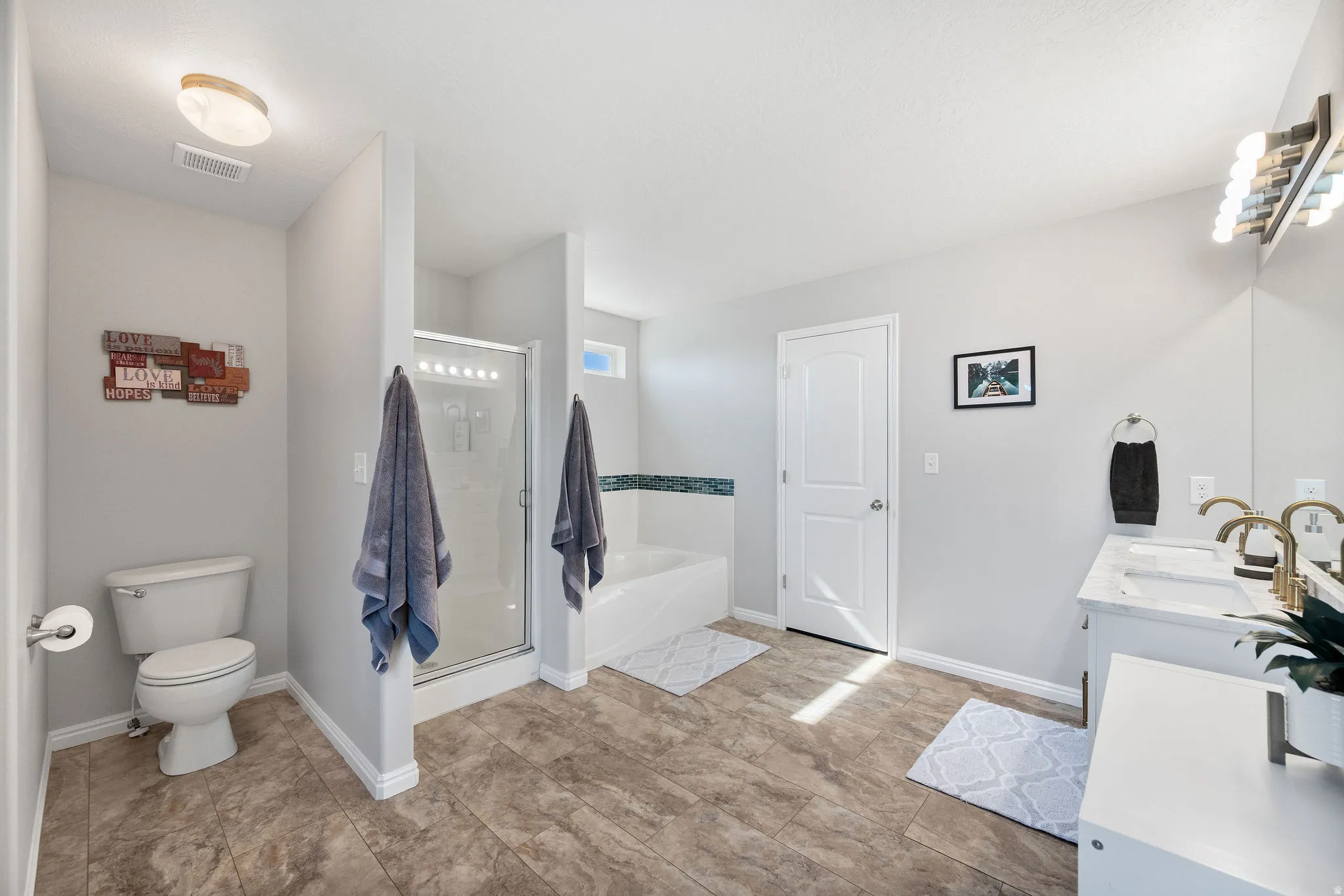 Spacious bathroom featuring wood-finish flooring, a glass-enclosed shower, a separate soaking tub with decorative tile accents, and a dual vanity with gold-tone fixtures