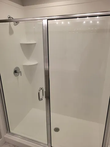 Enclosed shower featuring a clear glass door with chrome hardware, an integrated shower system with a rain shower head, dual corner shelves, and a white shower pan with a circular drain