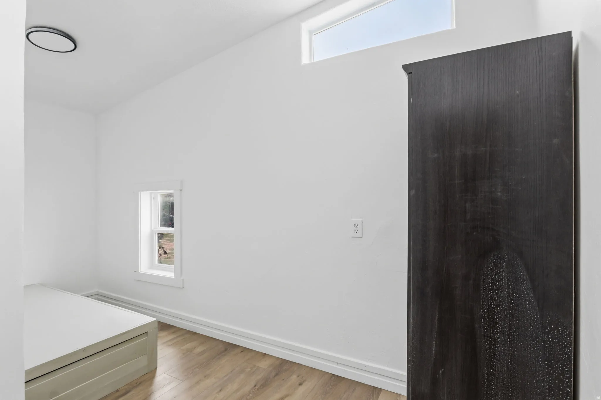 Minimalist interior featuring wood-finish flooring, a small framed window, and a high transom window providing ambient light