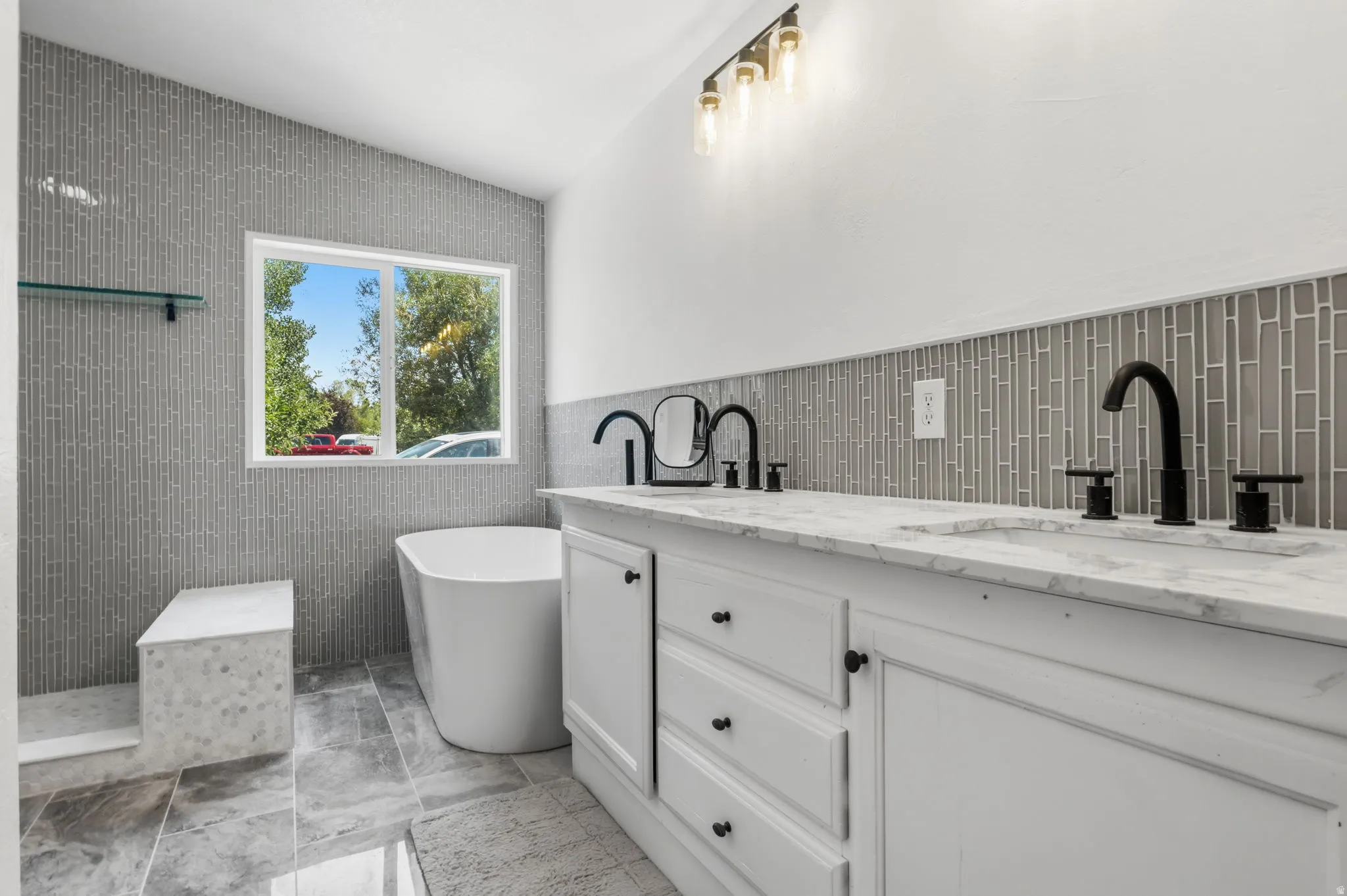 Contemporary bathroom featuring a freestanding tub, a dual vanity with marble-finish countertops, black hardware and fixtures, and a glass tile accent wall