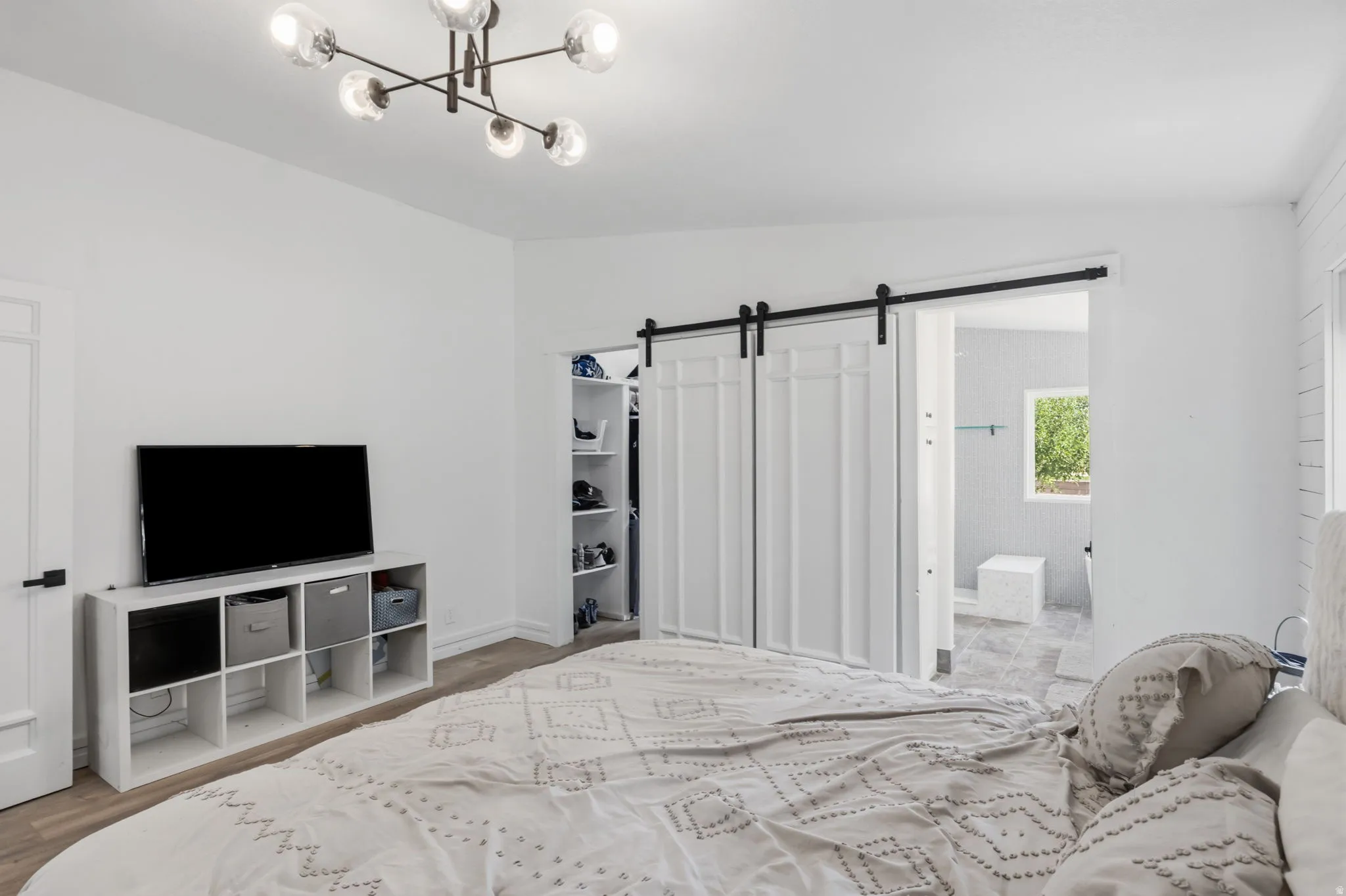 Bedroom featuring modern ceiling light fixture, white walls, wood-finish flooring, and a barn door leading to an adjacent bathroom