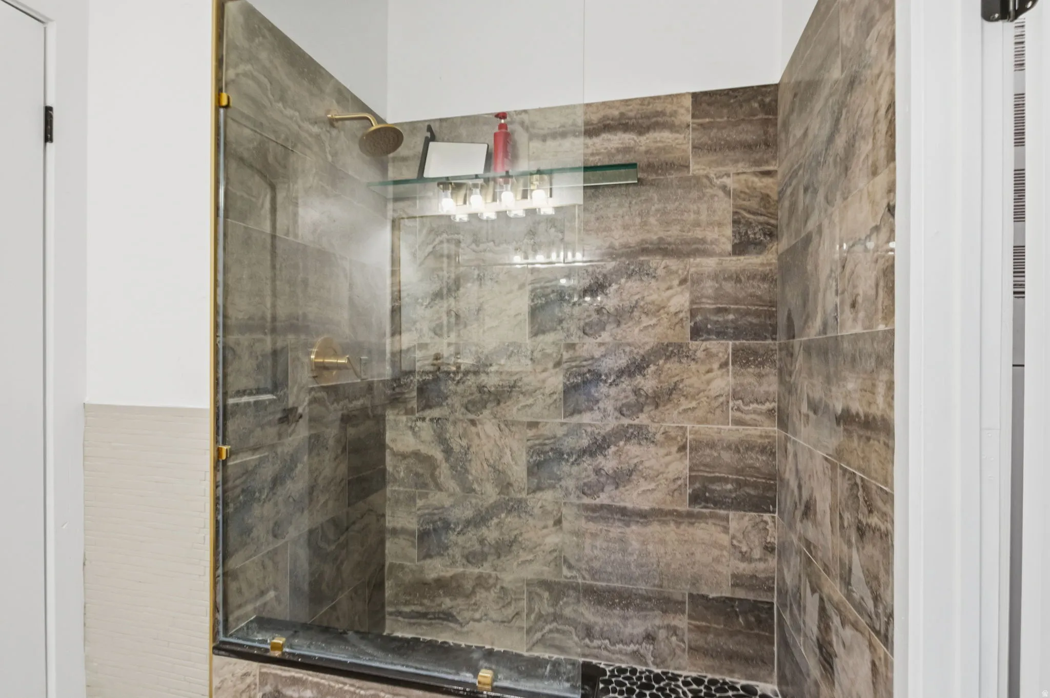 Walk-in shower featuring glass enclosure with brass-finish hardware, large format stone tile walls, a pebble-finish floor, and a built-in glass shelf