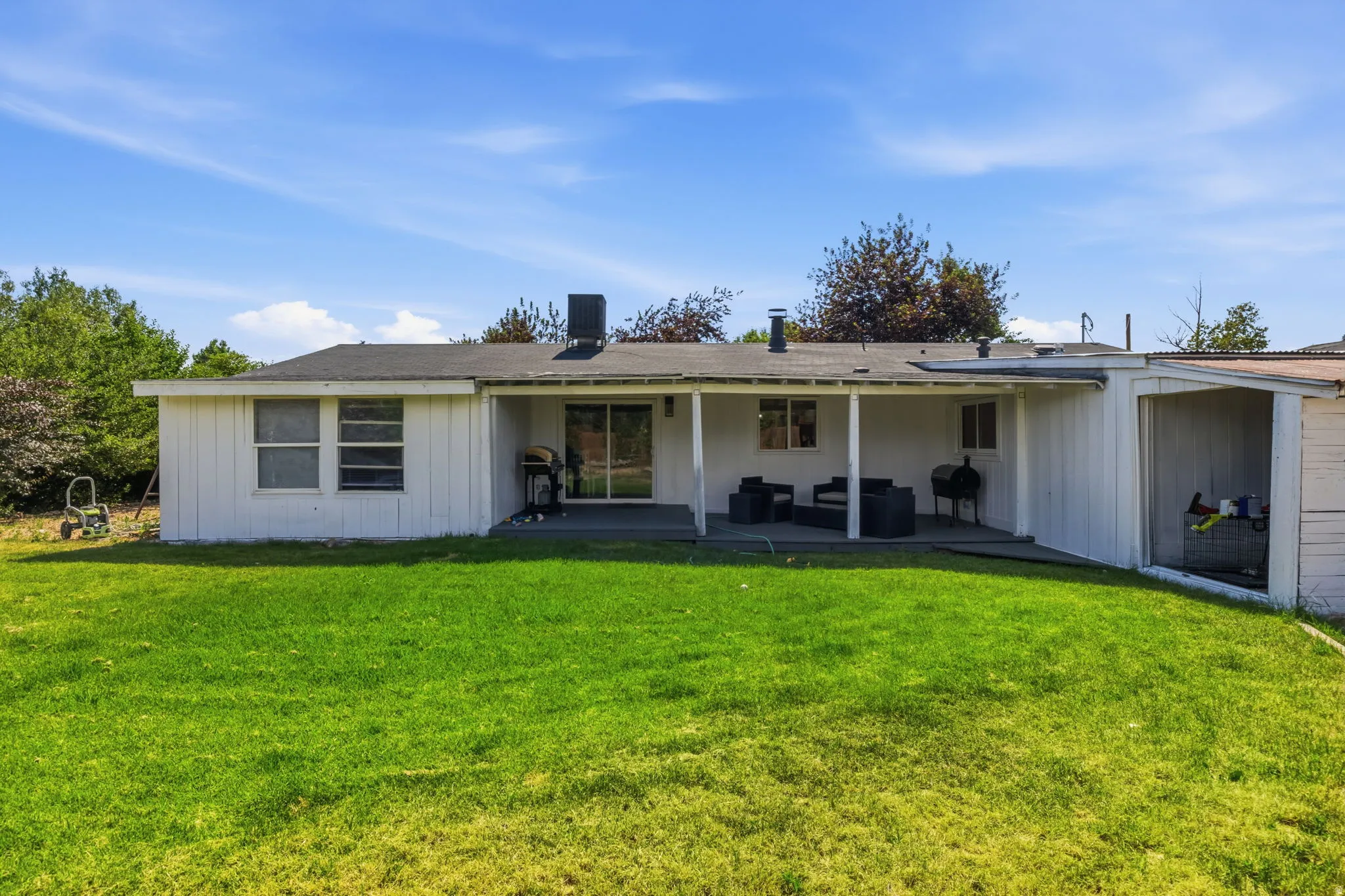 Spacious rear yard featuring a covered patio, sliding glass door access, white vertical siding, multiple windows, and an attached covered storage area