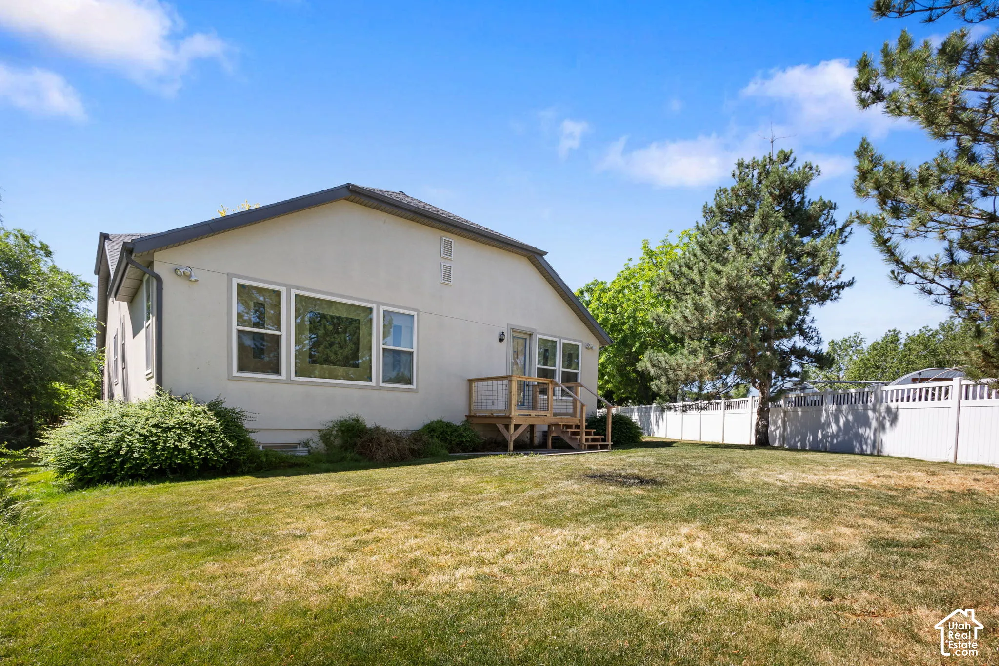 Rear exterior featuring a neutral stucco facade, wood deck with cable railing, mature trees, and a full-perimeter white vinyl fence