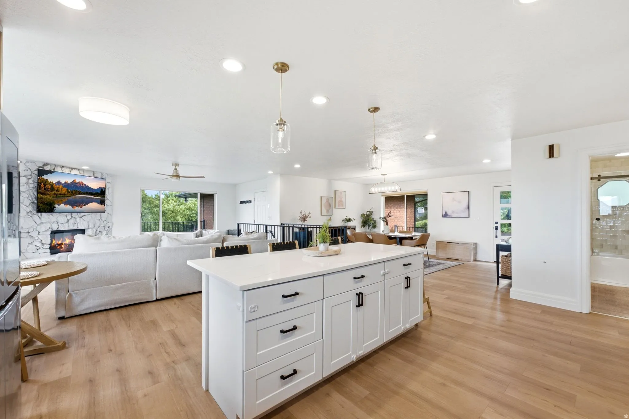 Open-concept living space featuring a central kitchen island with white cabinetry and a light-toned countertop, wood-finish flooring throughout, recessed lighting, and a stone-clad fireplace